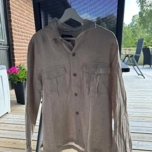 Beige linneskjorta från Riley - Snygg beige skjorta från Riley i luftigt linne. Skjortan har två stora bröstfickor med lock, klassisk krage och knäppning framtill. Perfekt för en chill och stilren look, passar till både jeans och shorts.