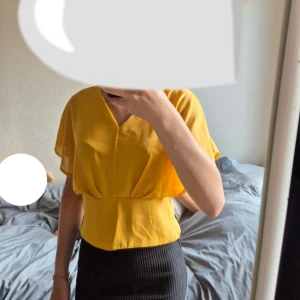 Gul blus med v-ringning och vida ärmar - Superfin gul blus med v-ringning både fram och bak samt en snygg detalj med ring i ryggen. Blusen har vida, luftiga ärmar och markerad midja som ger en cool siluett. Perfekt för dig som vill sticka ut med färg och stil.