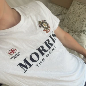 Vit t-shirt Morris The Gent - Vit t-shirt från Morris med stort tryck 'MORRIS THE GENT' på bröstet, broderad badge med lejon och brittiska flaggor samt siffran 68 i mörkblått på ärmen. Klassisk rund hals och korta ärmar. Perfekt för dig som gillar preppy stil.