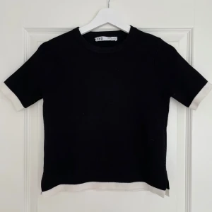 Svart stickad topp från Zara - Snygg svart stickad topp från Zara med kontrasterande vita kanter vid ärmslut och nederkant. Modellen har rund hals och korta ärmar, perfekt för en clean och stilren look. Passformen är normal och materialet känns mjukt och bekvämt. Inga defekter!