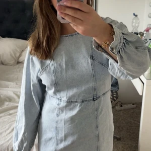 Ljusblå jeansklänning med långa ärmar - Säljer en snygg ljusblå jeansklänning i kort modell med markerade sömmar och långa ärmar. Klänningen har en rak passform och knappar vid ärmsluten. Perfekt för dig som gillar en avslappnad men trendig stil.