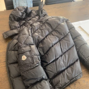 Svart Moncler dunjacka med huva - Säljer en svart Moncler dunjacka med huva och klassisk logga på ärmen. Jackan är quiltad, har dragkedja och en innerficka med serietidningsdetalj. Perfekt för kalla dagar och riktigt snygg streetstil.