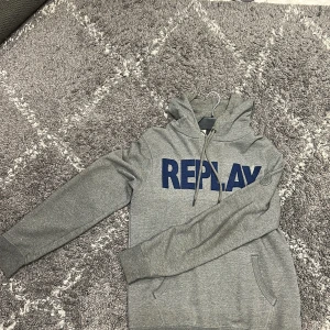 Grå hoodie från Replay med blå logga - Snygg grå hoodie från Replay med stor blå logga framtill. Tröjan har huva med snörning, känguruficka och långa ärmar. Perfekt för en chill och avslappnad stil. Tillverkad i mjuk bomullsmix som känns skön mot huden.