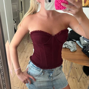 Vinröd korsett med snörning - Säljer en vinröd korsett med snygga volangdetaljer längs bysten och nederkanten. Toppen är axelbandslös och har snörning i ryggen för perfekt passform. Ger en riktigt cool och trendig look, perfekt att styla med jeans eller kjol.