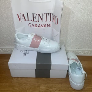 Valentino Garavani vita sneakers - Säljer ett par snygga sneakers från Valentino Garavani kopior. Vita med rosa rem. Storlek 39. Fint skick och kommer med   kartong + påse. Väldigt trendiga och bekväma.