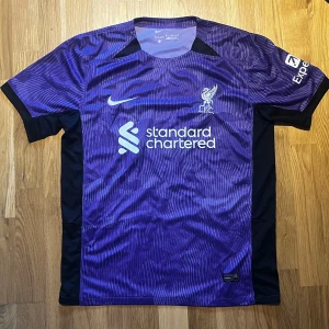 Liverpol Fotbollströja - Säljer en liverpol tröja från Nike med korta ärmar. Tröjan har ett subtilt randigt mönster och klubbens logga på bröstet. Perfekt för fotbollsfans som vill visa sitt stöd.