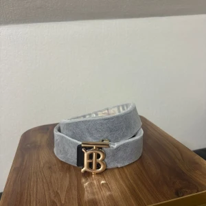 Burberry bälte med guldspänne - Tre snygga Burberry-bälten med ikoniskt TB-spänne i guld. Ett är ljusgrått i tyg, ett svart med diskret rutmönster och ett klassiskt beige med Burberry-rutor. Perfekt accessoar för att lyfta din outfit med lyxig känsla och stilren design.