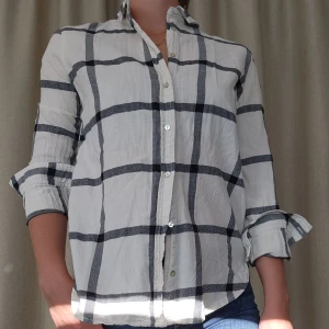 Vit och grå rutig skjorta - Snygg vit skjorta med stora grå och svarta rutor. Skjortan har knappar framtill, klassisk krage och långa ärmar som kan vikas upp. Perfekt till jeans för en avslappnad look.