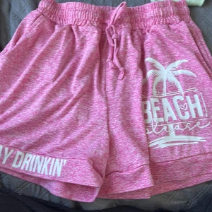 Rosa shorts med tryck och snören  - Snygga rosa shorts med vit text och palmtryck där det står 'BEACH please'. De har elastisk midja med snörning och är tillverkade i mjuk, stretchig polyesterblandning. Perfekta för sommardagar på stranden eller chill hemma.