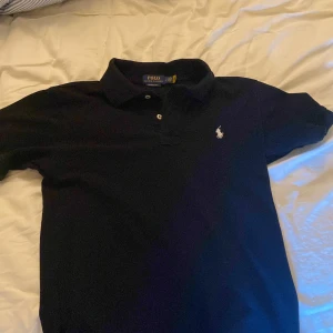 Svart pikétröja från Ralph Lauren - Klassisk svart pikétröja från Polo Ralph Lauren med vit broderad logga på bröstet. Tröjan har krage, korta ärmar och knäppning framtill. Perfekt för en clean och stilren look. Tillverkad i mjuk bomullg som känns skön mot huden. Inget håll eller skada