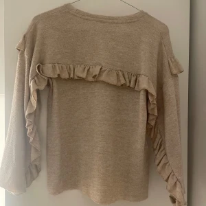 Beige tröja med volangdetaljer - Superfin beige tröja med volanger längs ärmarna och över bröstet. Mjuk och skön, perfekt för dig som gillar en söt och trendig look. Långärmad modell i stickad känsla, passar perfekt till jeans eller kjol.