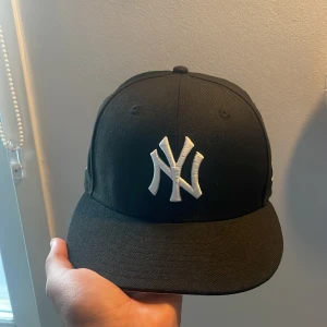 Svart New Era Yankees keps - Snygg svart snapback-keps från New Era med New York Yankees-logga broderad i vitt framtill. På sidan finns ett broderat World Series 1996-märke och New Era-loggan. Justerbar baktill med klassisk snapback. Perfekt för dig som gillar sportig streetstyle.