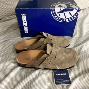 Birkenstock Boston  - Birkenstock sandaler i beige mocka med justerbart spänne i metall och klassisk korksula. Skorna har täckt tå och öppen häl, samt en mjuk innersula för extra komfort. Perfekta för en chill och avslappnad stil. Gjorde äkta mocka, Obs lnte 0rglnal 