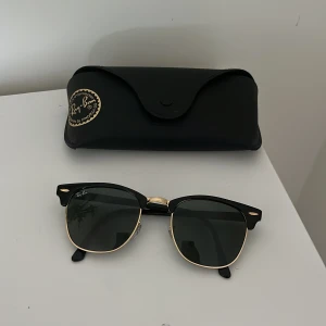 Ray-Ban Clubmaster solglasögon svart/guld - Snygga Ray-Ban Clubmaster solglasögon med svart ovandel och guldfärgade detaljer. Klassisk halvbåge och mörka glas. Kommer med Ray-Ban fodral. Perfekt accessoar för en cool och tidlös stil.