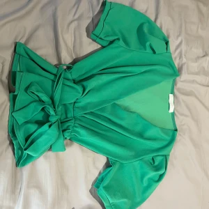 Grön omlottopp från Sisters Point - Snygg grön omlottopp från Sisters Point med korta ärmar och stor knytrosett i midjan. Toppens material är mjukt och lätt, perfekt för att skapa en cool och trendig look. Passar dig som gillar färg och vill sticka ut lite extra.