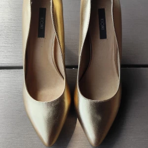 Guldiga pumps från Kiomi med klack - Snygga pumps i metallic guld från Kiomi med spetsig tå och hög klack. Skorna har en elegant form och är tillverkade i syntetmaterial med slät finish. Perfekta för dig som vill sticka ut med en glammig look.