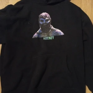 Svart hoodie från Hockey med tryck - Svart hoodie från Hockey med färgstarkt tryck på bröstet och texten 'HOCKEY' i grönt. Klassisk känguruficka framtill och huva. Perfekt för dig som gillar streetwear och vill sticka ut med en unik design.