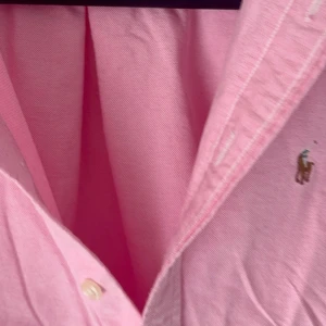 Rosa skjorta från Polo Ralph Lauren - Klassisk rosa skjorta från Polo Ralph Lauren med broderad logga på bröstet. Skjortan har lång ärm, knappar framtill och button down-krage. Tillverkad i mjuk bomull och har en normal passform. Perfekt för en stilren och avslappnad look.