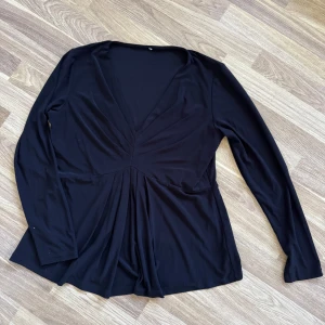 Svart v-ringad långärmad topp - Svart långärmad topp med djup v-ringning och snygga draperingar framtill. Modellen har en lösare passform nedtill och är tillverkad i ett mjukt, stretchigt material som känns skönt mot huden. Skulle gissa att det är storlek L, lappen är borta.