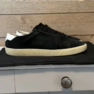 Svarta Saint Laurent sneakers i mocka - Snygga svarta sneakers från Saint Laurent med vit häl och vit sula. Skorna har klassisk låg profil, snörning och är tillverkade i mocka med detaljer i skinn. Perfekta för dig som gillar stilrena och exklusiva sneakers med en clean look.