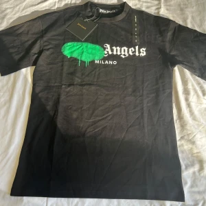 Palm Angels svart t-shirt Milano S - Svart t-shirt med grönt sprayat tryck och vit text 'Angels Milano' på bröstet. Klassisk passform, rund hals och korta ärmar. Grym streetwear-vibe och tillverkad i mjuk bomull. Perfekt för dig som gillar statement-plagg.
