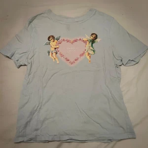 Ljusblå t-shirt med ängeltryck från H&M  - Ljusblå t-shirt från H&M Divided med korta ärmar och ett gulligt tryck av två änglar och ett hjärta med texten 'ANGELIC SOUL' framtrill. Storlek Xs