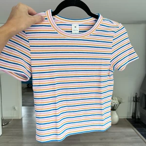 Randig croppad t-shirt - Superfin t-shirt med färgglada ränder i blått, rosa, orange och vitt. Inköpt på barnavdelningen. Klassisk rund halsringning och korta ärmar. Skön och stretchig i bomullsmix, perfekt för en chill och färgglad look. Önskar du veta exakta mått? Tveka inte att höra av dig! ALDRIG ANVÄND!