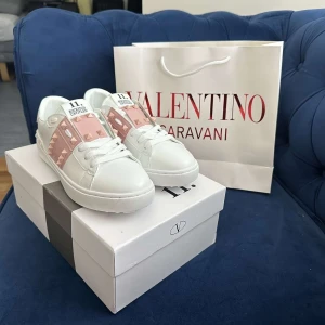 Valentino Rockstud sneakers vit/rosa - Snygga Valentino Rockstud sneakers i vitt skinn med ljusrosa band och ikoniska nitar. Klassisk rund tå, platt sula och vita snören. Märkeslogga på plösen och insidan. Perfekta för dig som vill sticka ut med exklusiva detaljer.