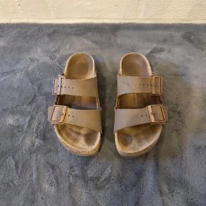 Bruna Birkenstock tofflor  - Klassiska Birkenstock sandaler i beige skinn med två breda remmar och stora metallspännen. Anatomisk fotbädd och platt sula för maximal komfort. Perfekta för varma dagar och enkel att matcha med allt.