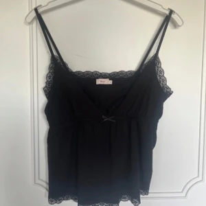 Svart spetslinnetopp från Nelly - Supersöt svart linnetopp från Nelly med tunna axelband, v-ringning och spetsdetaljer längs kanterna. Toppen har en liten rosett framtill och är lätt utsvängd nertill. Perfekt att styla med jeans eller kjol för en trendig look. Köpt för 260kr