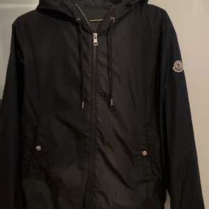 Svart vindjacka från Moncler - Svart vindjacka från Moncler med huva och dragsko, klassisk logga på ärmen och snygga silverdetaljer på dragkedja och knappar. Jackan är tillverkad i lätt polyester och har en clean, sportig look som passar perfekt till streetwear.