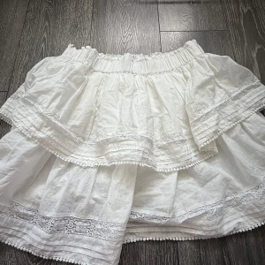 Vit volangkjol från Bik Bok - Supersöt vit kjol från Bik Bok med flera volanger och spetsdetaljer. Kjolen har bred resår i midjan och är perfekt för en somrig look. Lätt och luftig i bomull, med fina broderade kanter och romantisk känsla. Jättefint skick och söt kjol