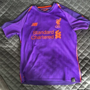 Äkta Liverpool Firmino 9 bortatröja lila - Liverpool FC bortatröja i lila med orange detaljer från New Balance. Tryck med Firmino 9 på ryggen och Premier League-märke på ärmen. Tröjan är kortärmad och tillverkad i lätt, ventilerande polyester. Perfekt för dig som älskar fotboll och vill sticka ut på planen.