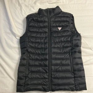 Svart dunväst från Guess - Snygg svart dunväst från Guess med quiltad design och dragkedja framtill. Västen har två fickor med dragkedja och en liten Guess-logga på bröstet. Perfekt att slänga över en hoodie eller tröja för extra värme.