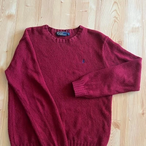 Vinröd stickad tröja Polo Ralph Lauren - Vinröd stickad tröja från Polo Ralph Lauren med rund halsringning och diskret blå logga broderad på bröstet. Klassisk modell med ribbade muddar vid ärmslut och nederkant. Perfekt för lager-på-lager och snygg till jeans.