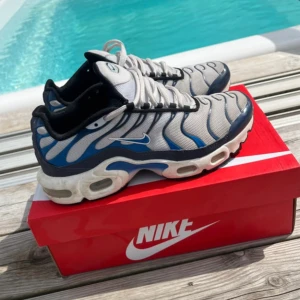 Nike Air Max Plus blå/vit sneakers - Nike Air Max Plus sneakers i blått, vitt och svart med ikoniska vågformade detaljer och synliga luftbubblor i sulan. Skorna har mesh-överdel, svarta och vita snören samt gult och rött TN-märke under sulan. Perfekta för dig som gillar streetwear och sportig stil.