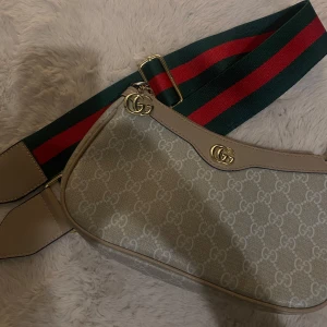 Beige axelväska  - Säljer en beige axelväska med klassiskt mönster över hela väskan. Väskan har detaljer i ljusbeige skinn och en bred axelrem i grönt och rött. Guldiga loggor och dragkedja upptill. Perfekt för dig som vill ha en ikonisk och trendig väska.