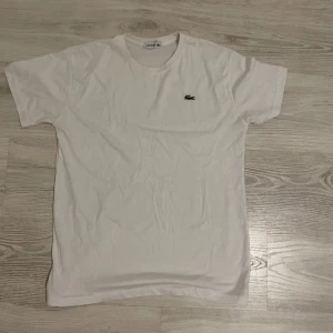Vit t-shirt från Lacoste i bomull - Vit klassisk t-shirt från Lacoste med den ikoniska gröna krokodilen broderad på bröstet. T-shirten är i mjuk bomull och har en enkel, stilren design med rund hals och korta ärmar. Perfekt för en clean och fräsch look.