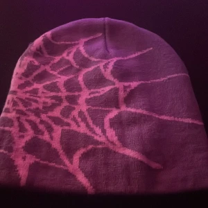 Lila mössa med spindelnätsmönster - Cool lila mössa med ett stort spindelnätsmönster i rosa över hela ytan. Perfekt för dig som vill sticka ut och gillar unika detaljer. Mössan är mjuk och stretchig, passar både till vardags och när du vill addera lite edge till din stil.