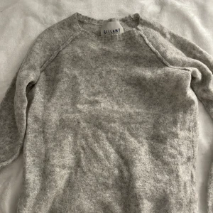 Ljusgrå stickad tröja från Bellamy - Mysig ljusgrå stickad tröja från Bellamy med rund halsringning och långa ärmar. Tröjan har en enkel och stilren design som passar perfekt till lager-på-lager. Materialet känns mjukt och värmande, perfekt för kyligare dagar.