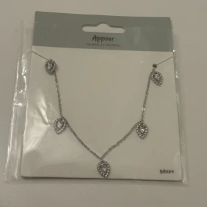 Silver halsband med droppar från Appear - Stilrent halsband i silverfärg från Appear med fem droppformade hängen, dekorerade med små glittrande stenar i hjärtformad design. Kedjan är tunn och smidig, perfekt för att ge en extra touch till din look. Säljs i originalförpackning. Oanvänt 