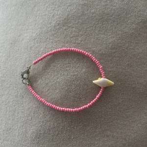 Rosa pärlarmband med snäckdetalj - Sött armband med små rosa pärlor och en unik snäckformad detalj i vitt och gult. Armbandet har ett silverfärgat lås och ger en somrig vibe. Perfekt accessoar för att liva upp din stil med lite färg och strandkänsla.                                                       Mått: 16 cm