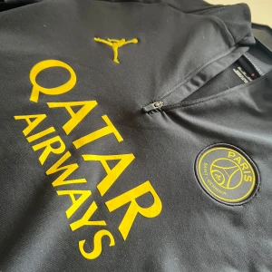 PSG x Jordan svart långärmad tröja - Svart långärmad PSG fotbollströja i slim fit från Jordan, Paris Saint-Germain-märke och Qatar Airways-tryck. Halvlång dragkedja framtill och tillverkad i ett sportigt, stretchigt material. Tight 