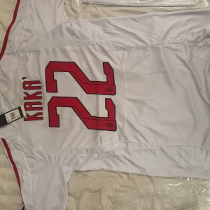 AC Milan Kaka #22 bortatröja Adidas - AC Milan bortatröja i vitt med röda och svarta detaljer, Adidas-logga och klubbmärke på bröstet. Kaka och nummer 22 på ryggen. Tillverkad i lätt och ventilerande polyester, klassisk passform och korta ärmar. Perfekt för dig som älskar fotboll och vill hylla en legend.