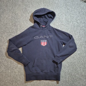 Mörkblå hoodie från GANT  - Snygg mörkblå hoodie från GANT i storlek XS. Tröjan har huva, magficka och ett broderat GANT-logo med sköld på bröstet. Perfekt för dig som gillar klassisk och sportig stil. Tillverkad i mjuk bomull för extra komfort.