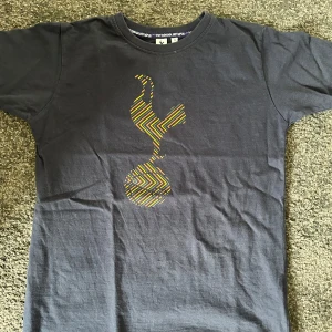 Tottenham T-shirt  - Visa ditt stöd för Tottenham Hotspur med denna stilrena mörkblå T-shirt – perfekt för både matchdag och vardag! Tillverkad i mjuk bomull med klubbens ikoniska tupp-logga framtill. Passar lika bra på läktaren som i soffan.
