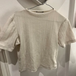 Ribbad beige t-shirt från Zara - ribbad/struktur t-shirt från Zara i en ljus beige färg. T-shirten har korta ärmar och en enkel, rak passform. 