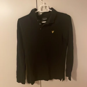 Svart pikétröja Lyle & Scott Junior - Svart långärmad pikétröja från Lyle & Scott Junior med klassisk krage och knäppning framtill. Tröjan har det ikoniska gula fågel-loggan broderad på bröstet. Perfekt för en clean och stilren kvällslook