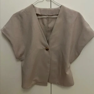 Beige topp med gulddetalj  - Säljer en stilren beige topp med v-ringning och korta ärmar. Toppen har en snygg guldig knapp framtill som detalj🤍Storlek S, köpt här på Plick men oanvänd båda av mig och förra ägaren☺️