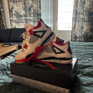 Nike Air Jordan 4 Retro Fire Red - Nike Air Jordan 4 Retro Fire Red med vit ovandel i skinn, svarta och röda detaljer samt meshpaneler. Klassisk siluett med synlig Air-sula, röda accenter och Jumpman-logga på plösen. Perfekta för dig som vill sticka ut med en ikonisk sneaker.
